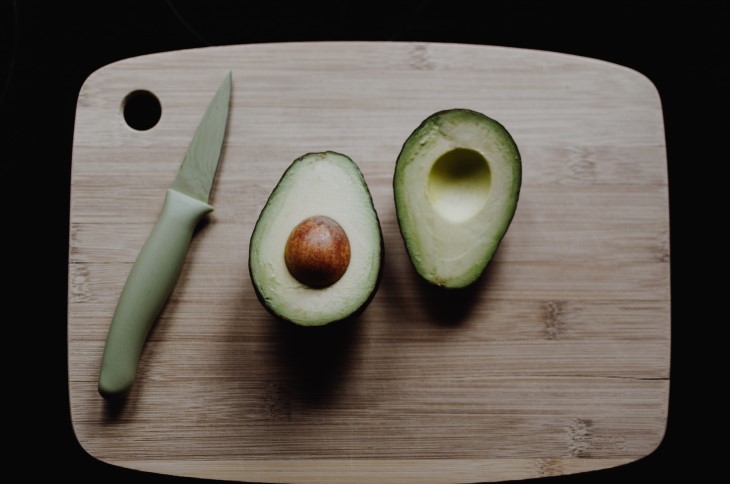 Avocado sits in half on a chopping board next to a knife