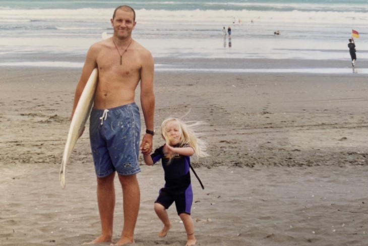 James holding a surf board and his daughter's hand on the beach