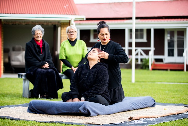 A Rongoā Māori practitioner using her healing methods on a woman while whānau watch on. 