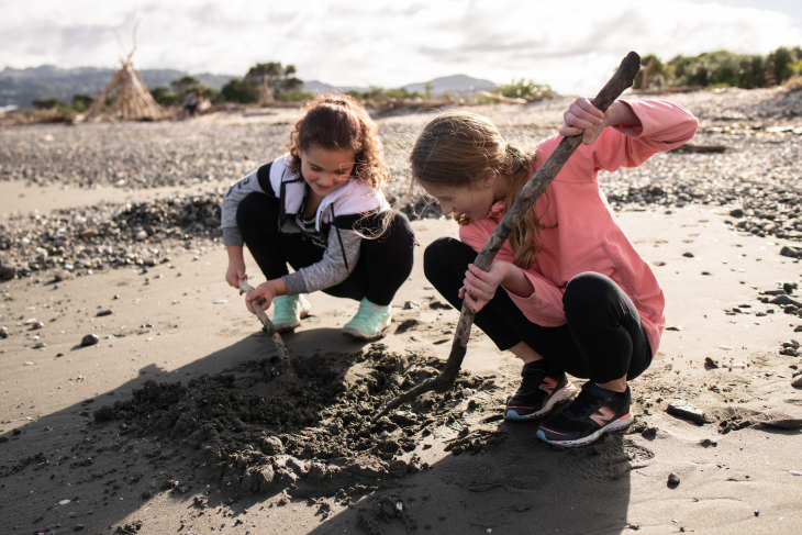 Two tamariki dig with some sticks on the beach