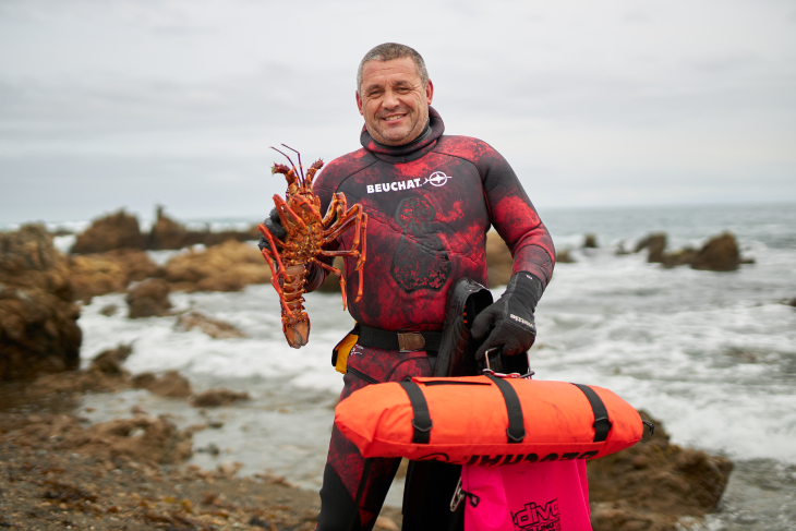 Todd Russell holds a crayfish and his dive gear beside the sea