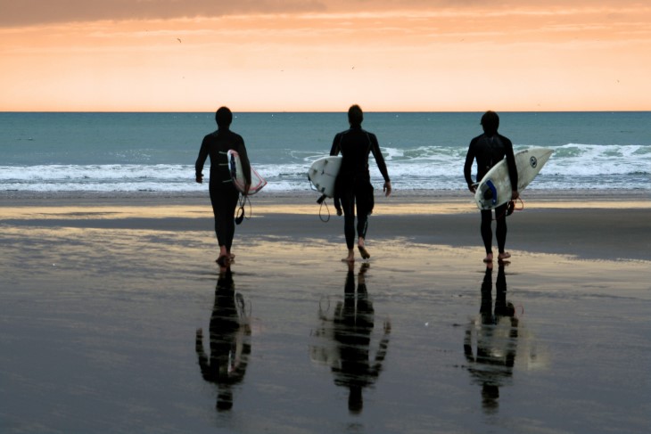 Three surfers head to the water in front of an orange sky
