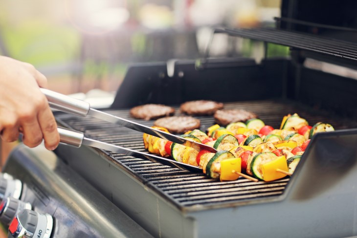 A close-up of a man's hand using tongs to cook vegetable kebabs on a barbeque. 