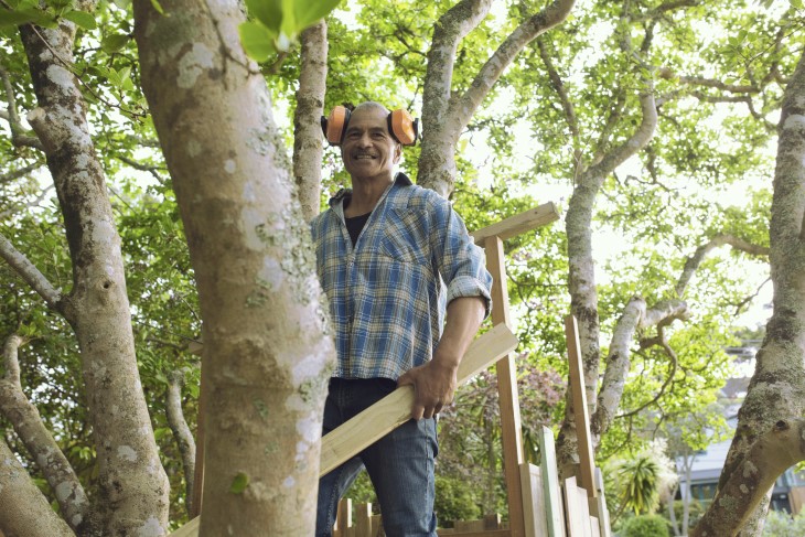 A man in his backyard carrying a plank of wood. 