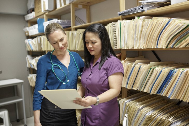 Doctor and nurse talk while looking at a file, and are standing in front of a wall of manilla folder