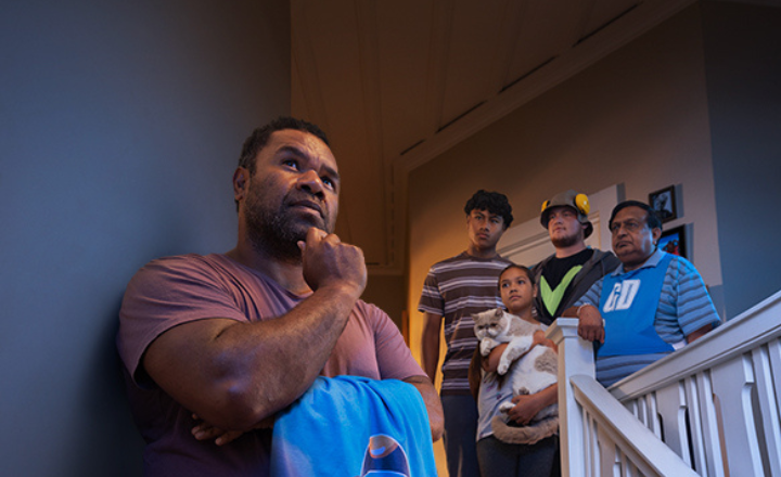 A father standing on stairs in his house doing the 'Have a hmmm' pose with family and friends behind