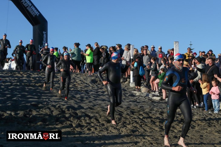 IronMāori competitors run along the beach in their wetsuits. 