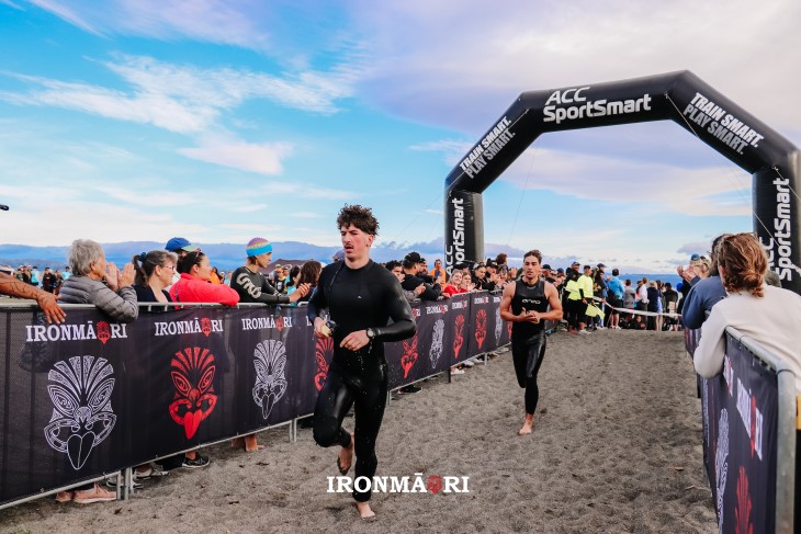 Two IronMāori competitors running up the beach with an ACC SportSmart sign behind them.