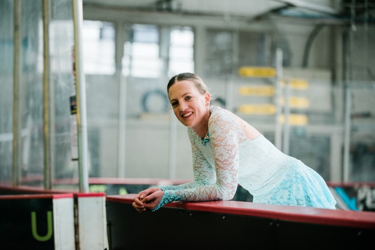 Lydia leans over the side of an ice skating rink in her blue performance outfit, and smiles to the camera