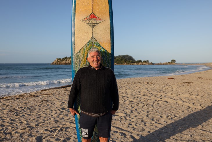 Michael stands with his surfboard on the beach