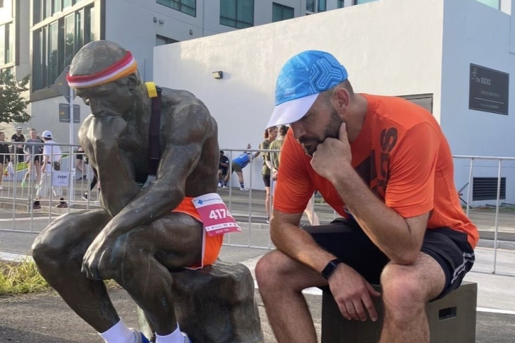 A man in running attire sits next to a copy of Rodin's scultpure 'The Thinker' which has also been adorned in sports clothing