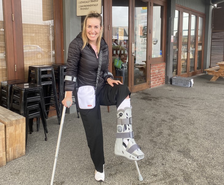 Alicia Mabey standing on her crutches with her moonboot and smiling.
