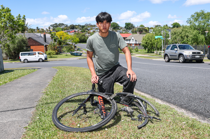 Borynt Em Ream kneeling down next to his broken bike on a grass verge next to the road.