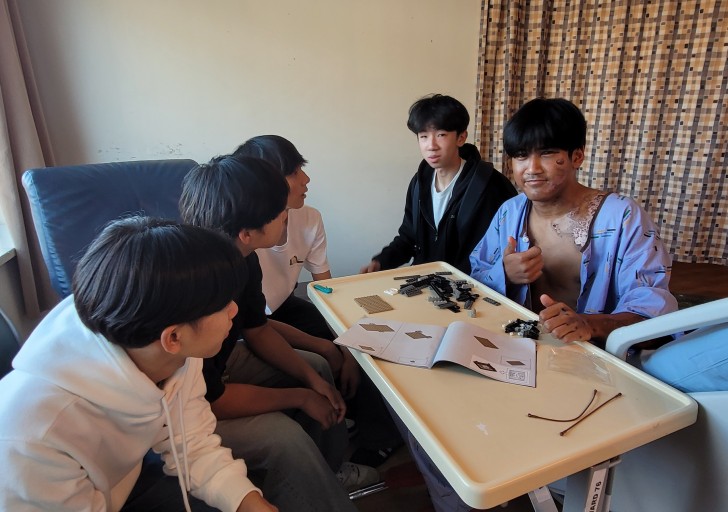 Borynt Em Ream sitting in hospital with several of his friends and giving the thumbs up.