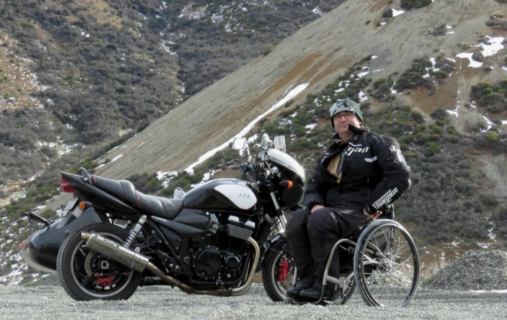 brett ladbrook 1 Brett Ladbrook sitting in his wheelchair in front of his motorbike with a mountain behind him.