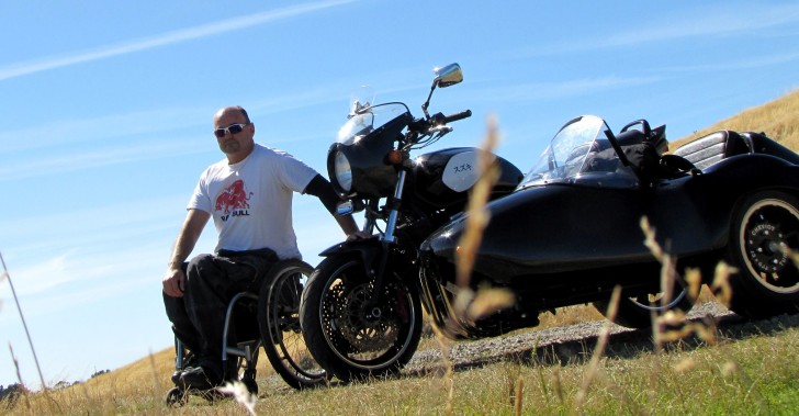 Brett Ladbrook sitting in his wheelchair next to his motorbike in front of a blue sky.