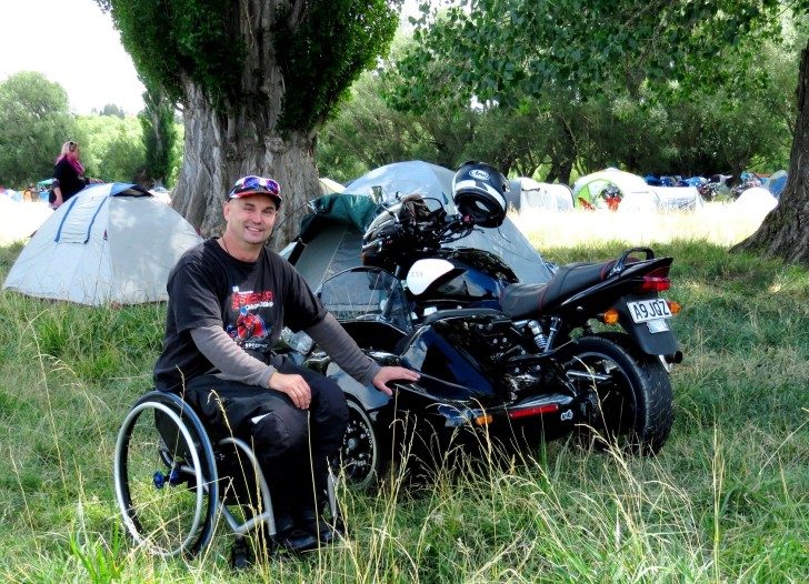 Brett Ladbrook sitting in his wheelchair next to his motorbike in a camp ground.
