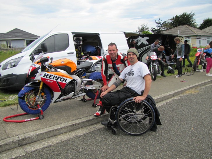 Brett Ladbrook sitting in his wheelchair next to a motorbike with another man wearing motorcycle gear.