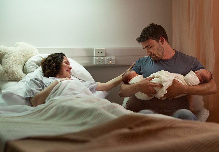 A man proudly holding newborn twins in his arms while his partner lies in a bed next to him.
