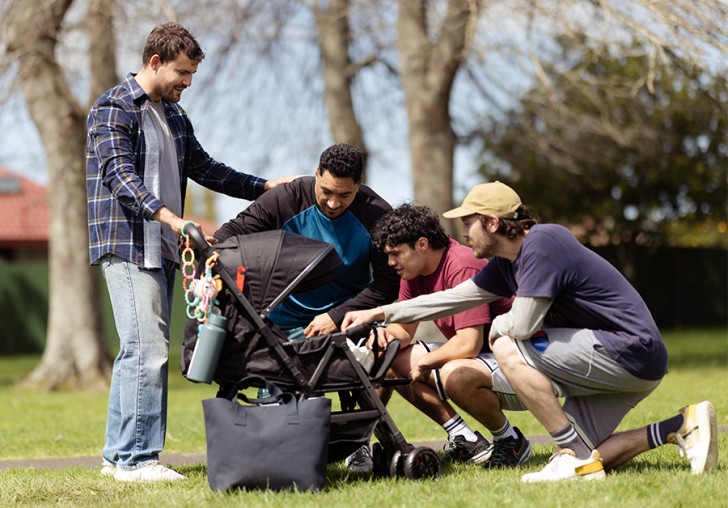A man holding a pram in a park while three of his friends look into the pram to see the baby.