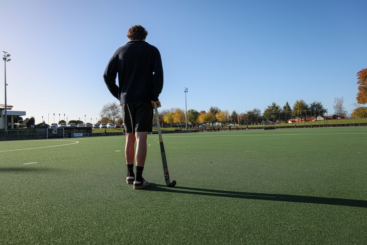 Campbell Gray standing on a hockey turf and leaning on his hockey stick, facing away from the camera.