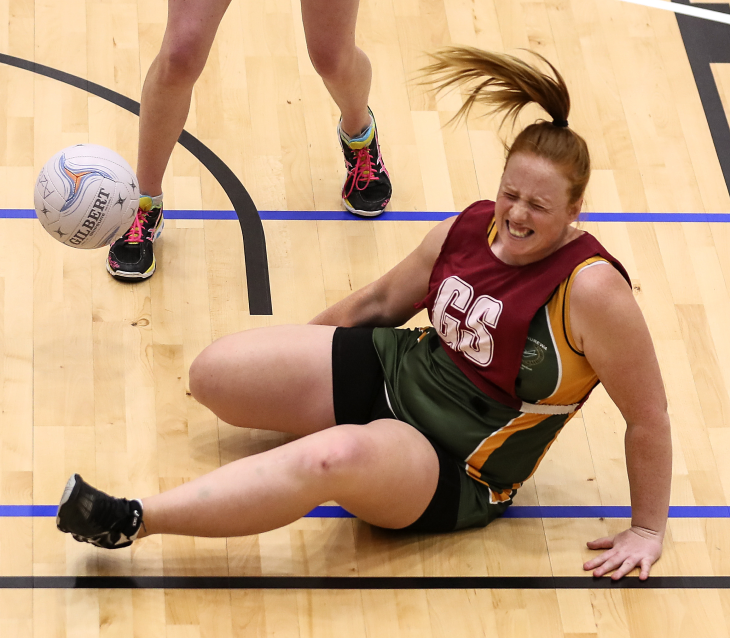 A netball player sitting on the court grimacing after suffering an injury.