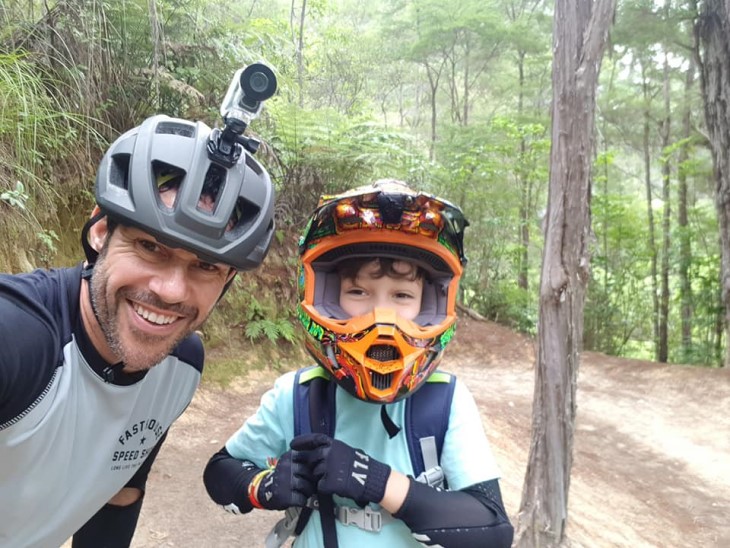 A selfie photo of Craig Harrington with his son, both are wearing helmets and standing on a mountain bike path.