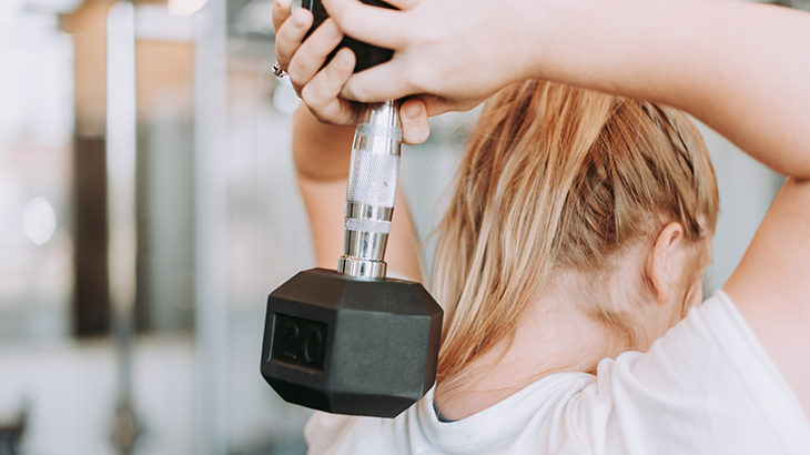 A woman holding a dumbbell weight behind her head at the gym.