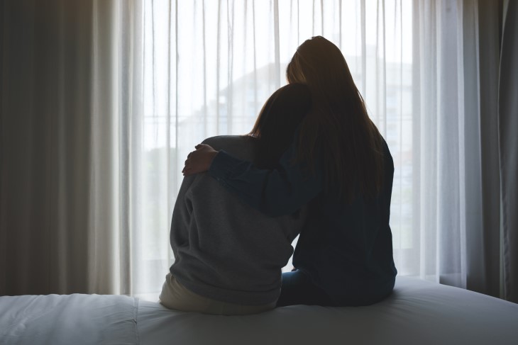 A silhouette shot taken from behind of a woman sitting on a bed with a child and comforting them.