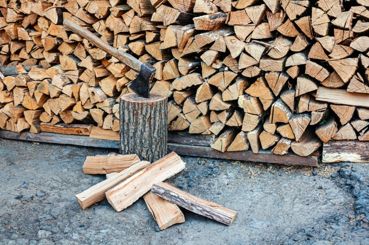 An axe sitting upright in a log of wood with a pile of chopped firewood behind.