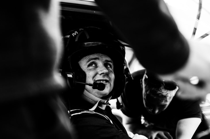 A black and white photo of James Dunlop sitting in his car with a helmet on and looking up at someone talking to him.