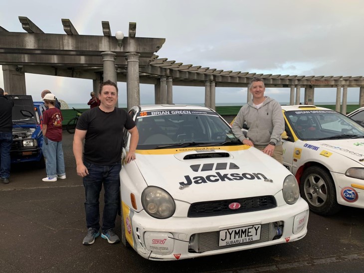 James Dunlop standing next to his rally car with another man standing on the other side, both are smiling at the camera. 