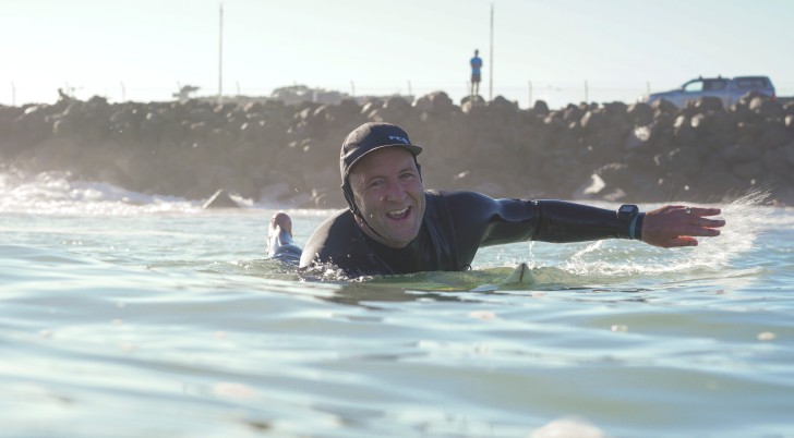 James Whitaker paddling on his surfboard and smiling.