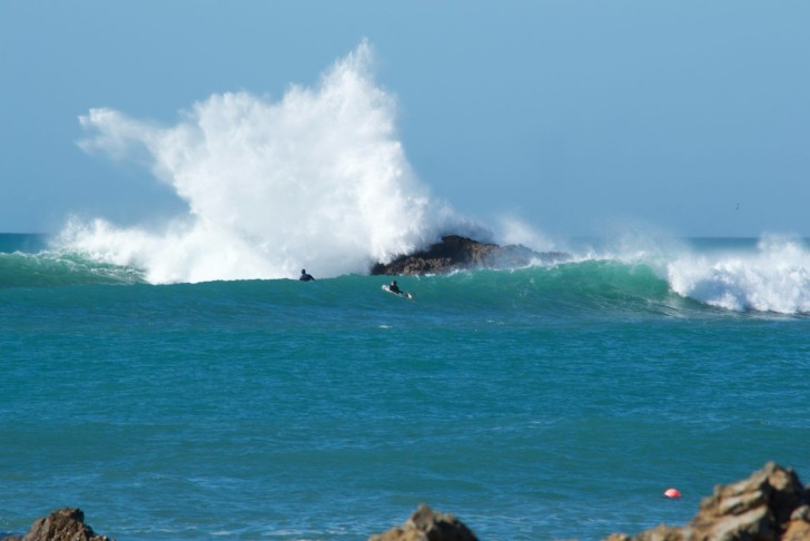 Two surfers in the water near a wave crashing onto a rock.