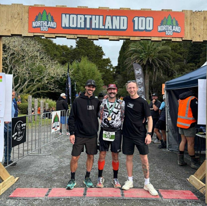 Mark Baker standing at the finish line with two friends after completing a long-distance running event.