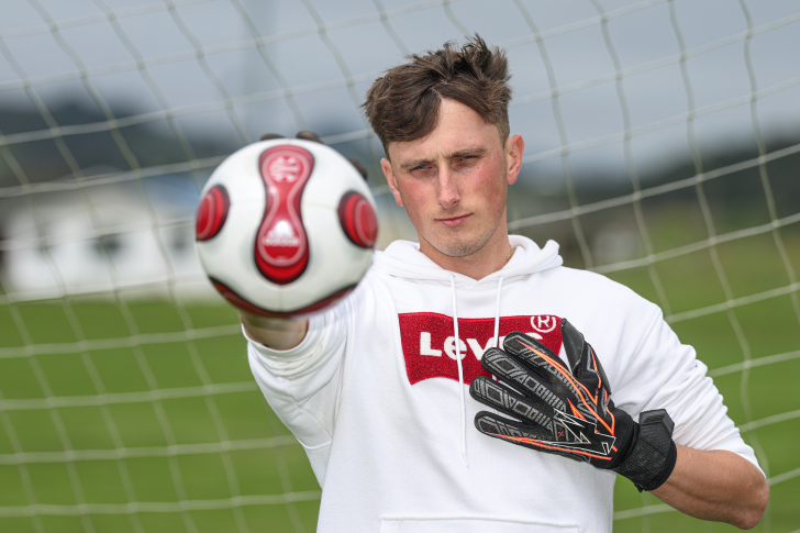 Matt Burchell wearing football goalkeeping gloves and holding the ball out towards the camera with his right arm.
