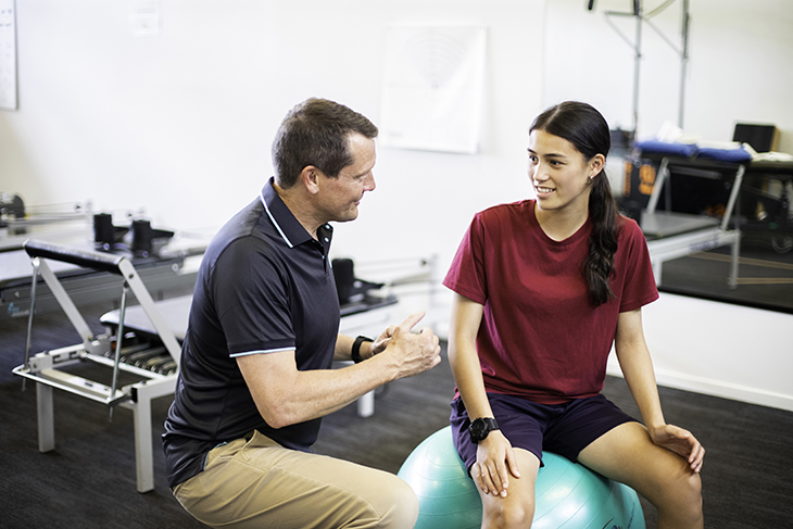 A male physio talking to a female patient, who is sitting on a Swiss ball in the gym. 