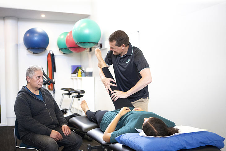 A young woman receiving physio treatment on her leg while an older man looks on.