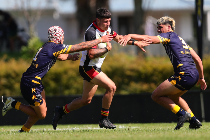 A young male rugby league player running with the ball between two players trying to tackle him.
