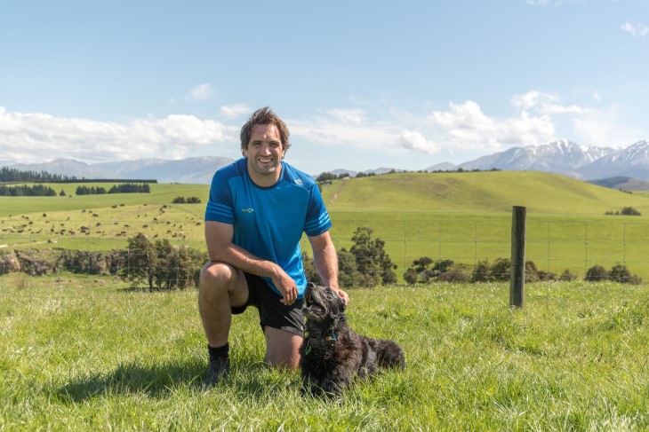 All Black Sam Whitelock kneeling down with a dog on a farm and smiling. 