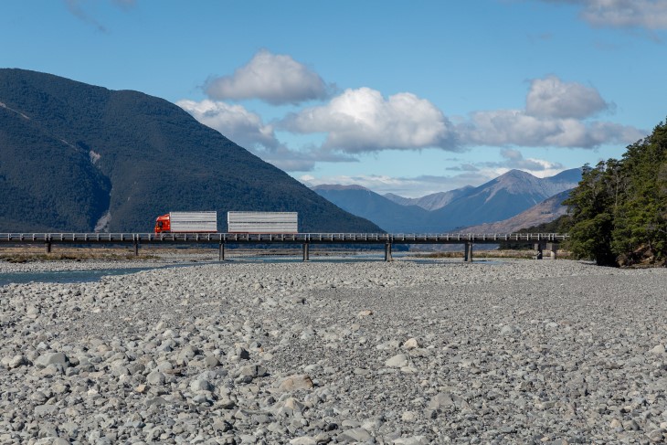 A large truck driving over a bridge across a river in front of mountains.