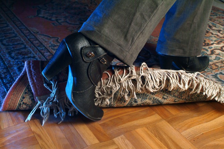 A close-up shot showing a woman's lower leg wearing a high-heeled shoe and tripping over a rug on a wooden floor.