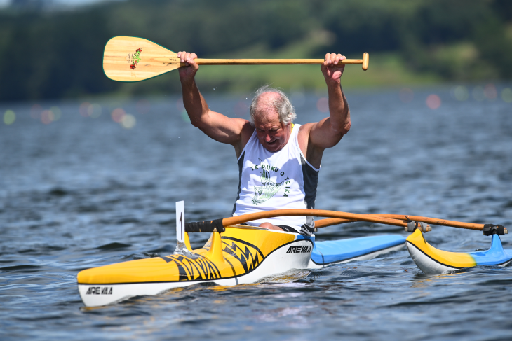 An older man holding his paddle above his head in an outrigger canoe after a waka ama race. 