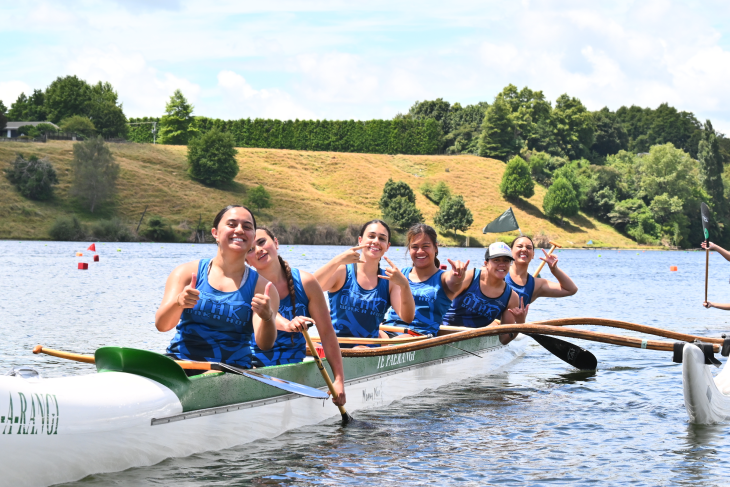 A group of young women posing for the camera in an outrigger canoe on a lake.