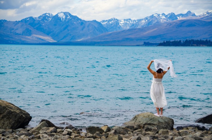 A bride stands on a lakeside in front of stunning mountains. 